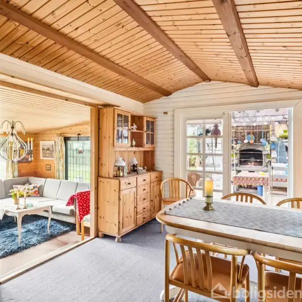 Træbord omgivet af træstole står i en stue med trægulve og loft. Døren fører til en terrasse med udendørs siddepladser. Hylder og glasdør skaber en hyggelig atmosfære.