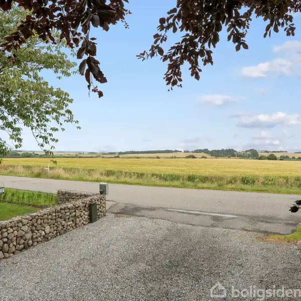 Grusbelagt indkørsel med stendige, træer hænger over. Rolig vej krydser foran med åbne marker i baggrunden under en næsten skyfri blå himmel.