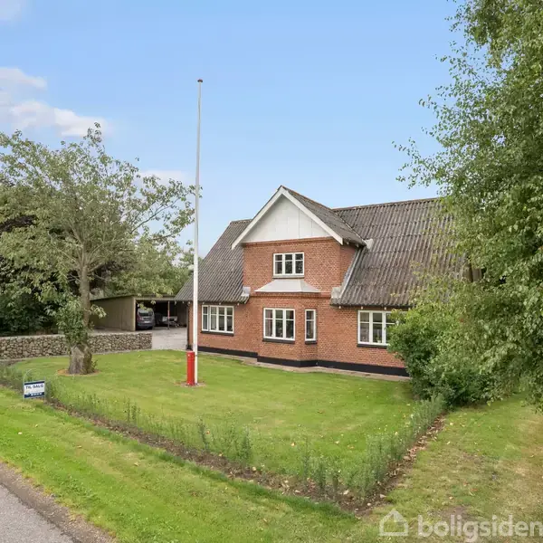 Et rødt murstenshus står stille på en grøn plæne med en flagstang foran, omgivet af træer og en grusvej. En "til salg"-skilt markerer ejendommen.