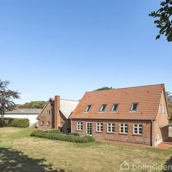 En rød murstenshus med tagvinduer står på en grusvej. Huset har en stor træ i haven under en klar blå himmel.