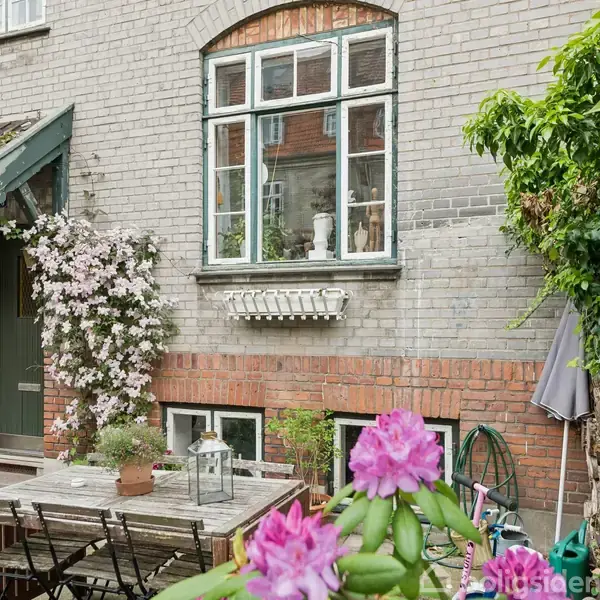 En træbord med stole står i en gårdhave ved en murstensbygning. Røde og lyserøde blomster i forgrunden, og klematis klatrer op ad væggen ved siden af en grøn dør.