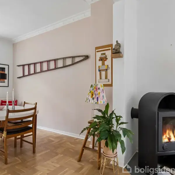 En spisestue med et rundt bord og fire stole. Et tændt brændeovn står til højre. Vægdekorationer inkluderer en stige. Grønne planter og en farverig lampe omkranser rummet.