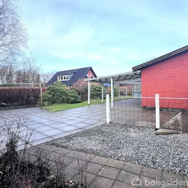 Rød murstensbygning står i forgrunden med en integreret carport. Omgivet af flisebelagt terrasse og småsten. Træer og buske i baggrunden under en klar himmel, typisk villavejscene.
