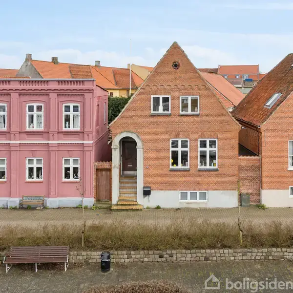To huse står ved siden af hinanden; det ene er pink med hvide vinduer og dekorative detaljer, det andet er rød mursten med asymmetrisk tag. Forgården har en bænk og buske.