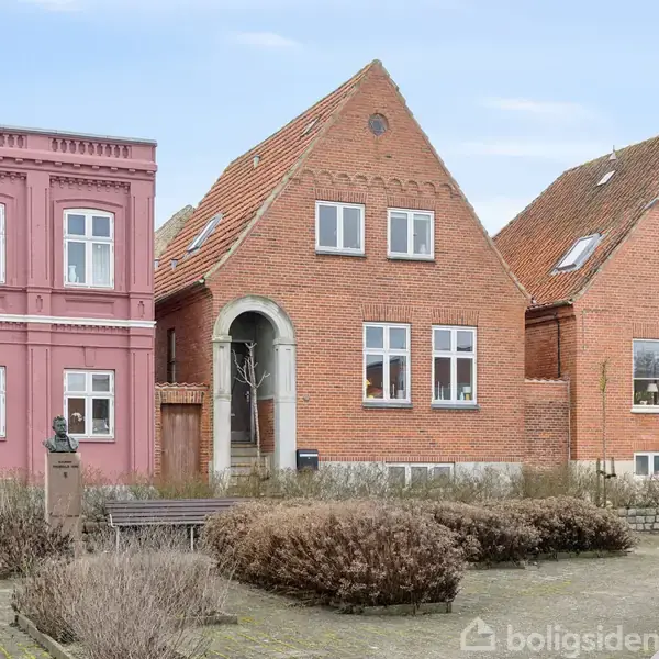 Rødt murstenshus med spidst tag og hvide vinduer. Huset har en bueformet indgang. Til venstre ses et lyserødt hus. Foran er en gårdhave med buske og en buste på piedestal.