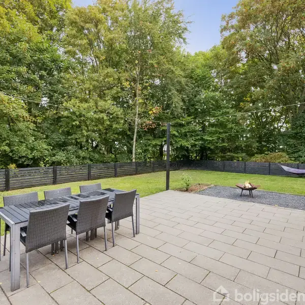 Terrassebord med seks stole står på en flisebelagt terrasse. I baggrunden er en græsplæne og tætte træer. En bålplads og hængekøje er placeret nær et mørkt træhegn.