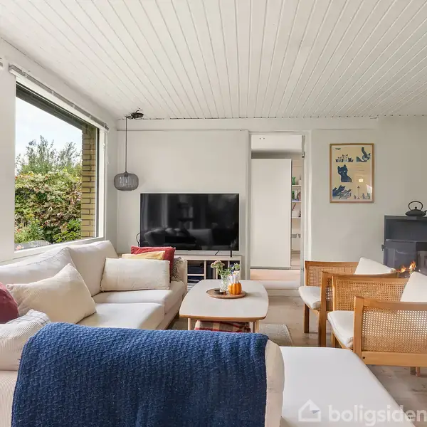 En stue med en stor, hvid hjørnesofa dækket med farverige puder. Foran sofaen står et sofabord med dekorationer. Kort til højre ses en brændeovn og en TV-skærm. Udenfor vises en frodig have gennem store vinduer.