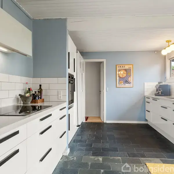 Et moderne køkken med hvide skabe og sorte håndtag. Detaljer omfatter en kogezone med krydderier, flisevægge, træskilt og blå vægge. Vinduer slipper naturligt lys ind.