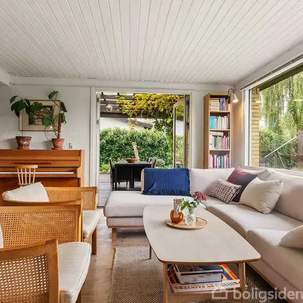 Stue med hvid sofa og træmøbler, hvor en klaver står ved siden af en bogreol. Store vinduer giver udsigt til frodig have med terrasse og grøn beplantning udenfor.