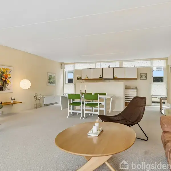 Stue med grøn lænestol og brun sofa står på et lyst tæppe. Spisebord med grønne stole og dekorative lamper i et åbent rum. Veggene er dekoreret med kunst.