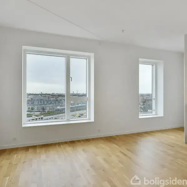 Rum med lyse trægulve og to store vinduer indsigt over byen, hvilket bringer naturligt lys ind. Køkkenet er synligt i højre side med hvide skabe.