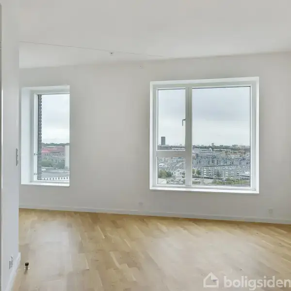 Et lyst rum med trægulv har to store vinduer, der afslører en udsigt over en by. Væggene er hvide, og der står en åben dør til venstre.