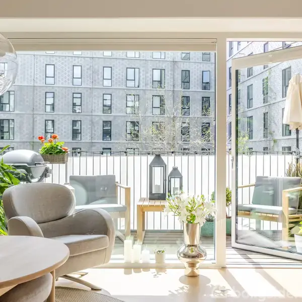 Lys stue med en rund stol og bord, fører ud til balkon med planter og møbler. Store vinduer viser udsigt til en moderne bygning. Glasdør står åben. Orkidé på bordet.