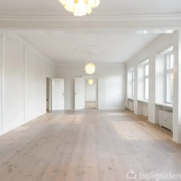 En stor, lys stue med trægulv strækker sig over det åbne rum. Loftlamper hænger ned. Store vinduer langs højre væg giver naturligt lys.