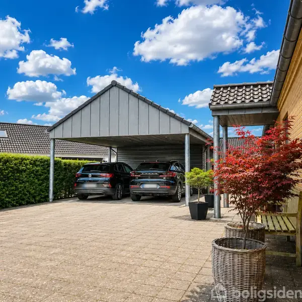 Carporten huser to parkerede biler ved siden af en gul murstenshus. Foran er belagt fliseplads med planteskjulere og en rød busk. Himlen er klar med spredte skyer.