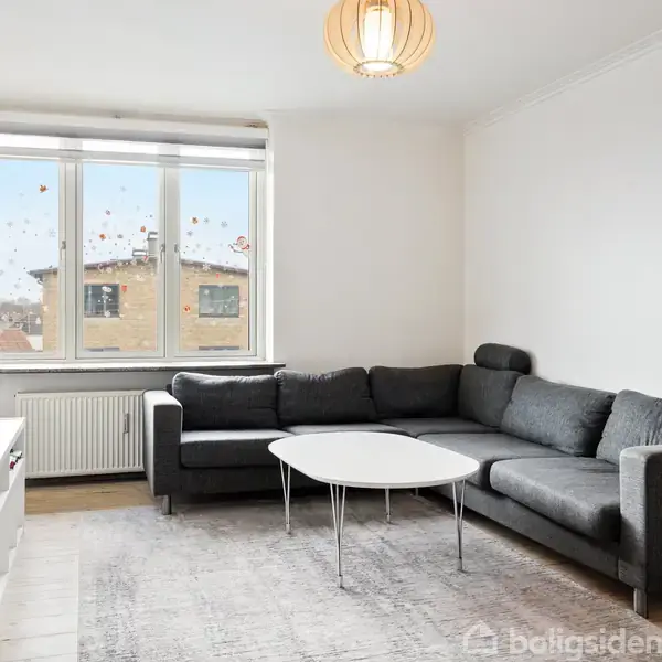 Grå hjørnesofa står i hjørnet under et stort vindue dekoreret med klistermærker. Et rundt hvidt sofabord står foran sofaen, med en lampe hængende ovenover.