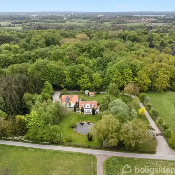 Et stort hus med rødt tag omgivet af frodig, tæt skov. Foran huset er en lille dam. En lang indkørsel fører gennem grønne marker til ejendommen.