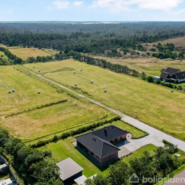 Hus med mørkt tag står stille ved en landevej omgivet af åbne marker. Rundt omkring ligger campingvogne langs en grusvej, og skove strækker sig i baggrunden.