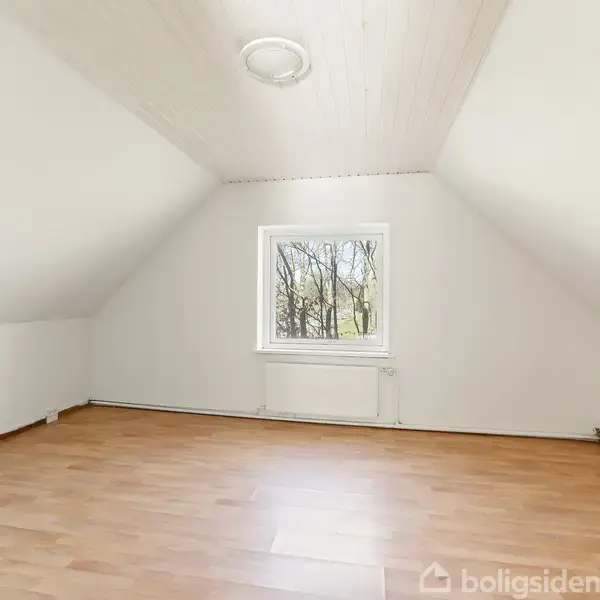 Et lyst rum med skrå vægge og et trægulv. Rummet har et vindue med udsigt til træer udenfor. En loftlampe er placeret centralt i loftet.