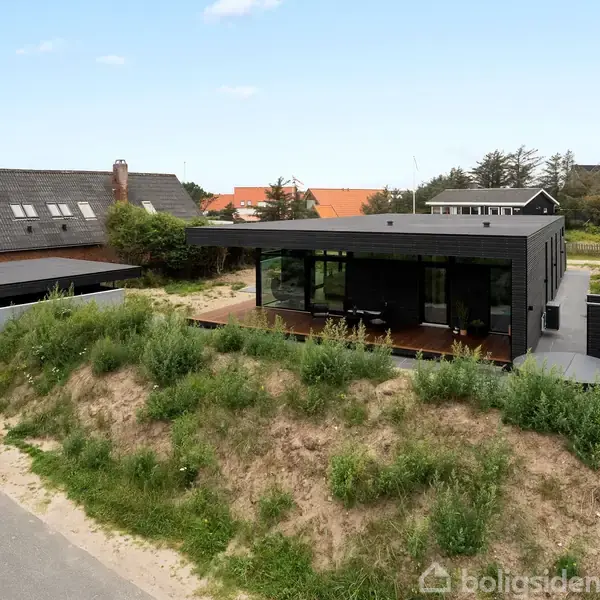 Et moderne sort hus står stille på en græsklædt dune. I baggrunden er der nabolag med flere huse under en blå himmel med få skyer.