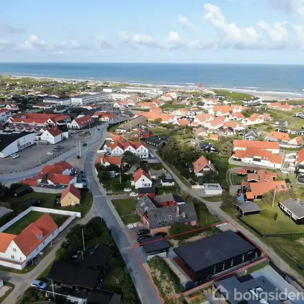 En kystby med røde og hvide tage spredt over et grønt landskab. Husene ligger langs snirklede veje med havet i baggrunden under en klar, blå himmel.