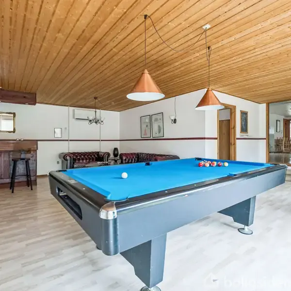 Poolbord med blå filt står ubenyttet i et lyst rum med trægulve og træloft. Bar med barstole og sofa ses i baggrunden, darttavle på væggen.