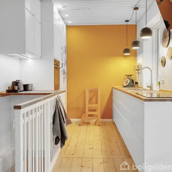 Et køkken med hvide skabe og træbordplader, hænger lamper over bordet. Mosaikfliser pryder væggen, og en orange væg giver farve til rummet. En træstol står bagerst.