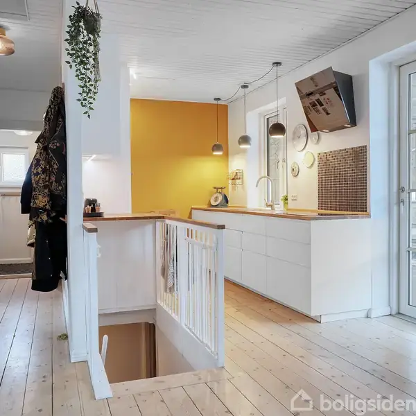 Et lyst køkken med hvide skabe, en gul accentvæg og træbordplade. En trappe leder nedad fra gangen. Store vinduer giver lys, og der hænger jakker på væggen.