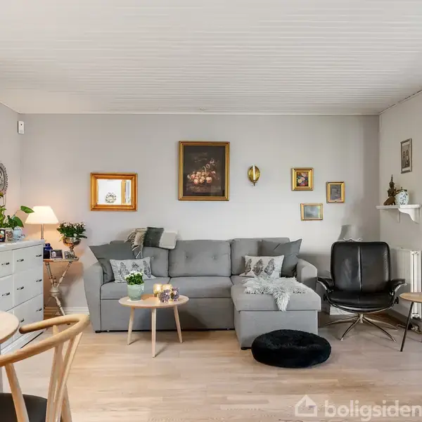 En stue med en grå sofa og sort lænestol står ved siden af et rundt bord. Væggene er dekoreret med flere malerier, og der er et hvidt skab og en pejs.