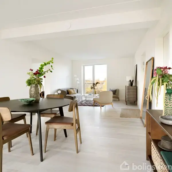 Rundt spisebord med træstole, står i et lyst rum. Der er grønne planter som dekoration og en sofa i baggrunden. Store vinduer giver udsigt til altan udenfor.