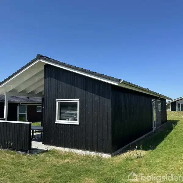Et sort, træbeklædt sommerhus med overdækket terrasse står på en græsplæne under en blå himmel. Store vinduer pryder husets facade, og der er et nærliggende bygning i baggrunden.