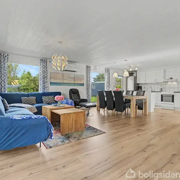 Blå sofa i hjørnet med puder, står i en stue ved siden af et træbord. Pejs til højre. Køkken med hvide skabe og spisebord i baggrunden. Store vinduer slipper lys ind.