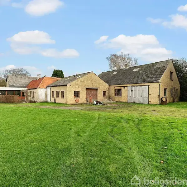 Et ældre bondehus i lysegule mursten med rødt og gråt tegltag står på en stor, velholdt græsplæne. I baggrunden ses flere bygninger og træer under en blå, skyet himmel.