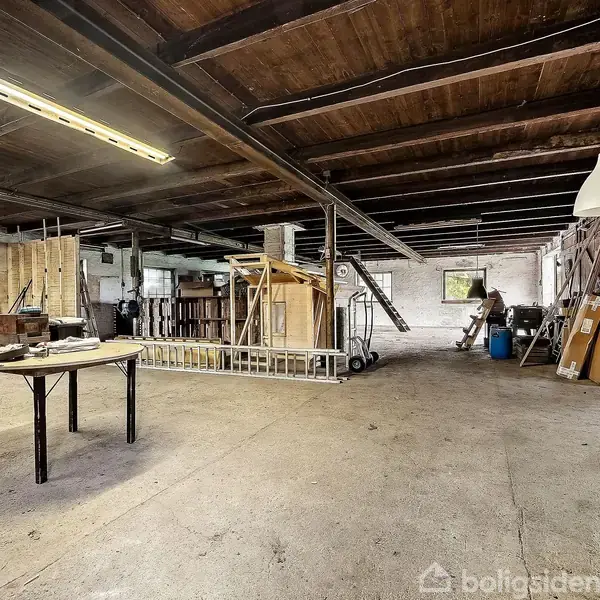 Værksted med træloft, belysning og bord til håndværk. Omgivet af værktøj, materialer og affaldscontainere i et rummeligt, industrielt rum. Vindue i baggrunden til venstre.