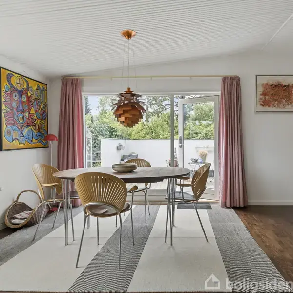 Et spisebord med træstole står på en stribet tæppe. En kobberfarvet loftslampe hænger over bordet. Store vinduer med lyserøde gardiner viser en grøn udsigt udenfor.