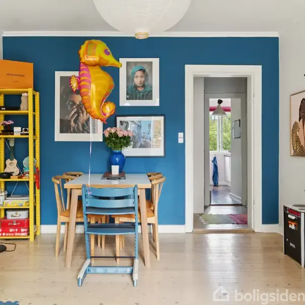 En spisebord med en blå vase og en ballon formet som en søhest står i stuen. Værelset har en blå væg, kunst på væggene, og bogreoler omkring.