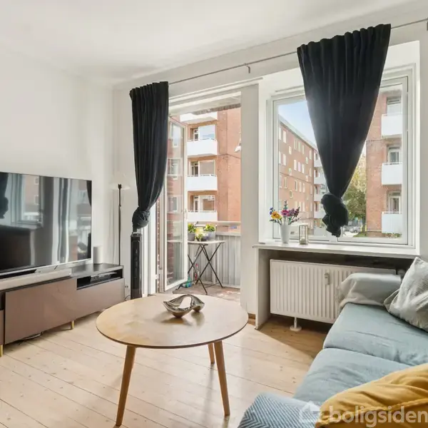En stue med sofa og sofabord foran et stort TV. Store vinduer med sorte gardiner åbner op til en balkon med udsigt til murstensbygninger.
