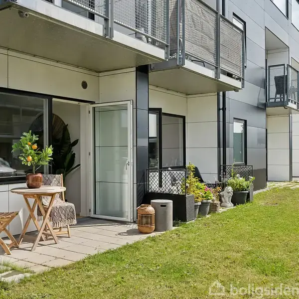 Terrassemøbler står på en lille terrasse foran en moderne bygning. Et bord med blomster i en krukke og to stole danner en hyggelig siddeplads. Græsplæne omgiver området.