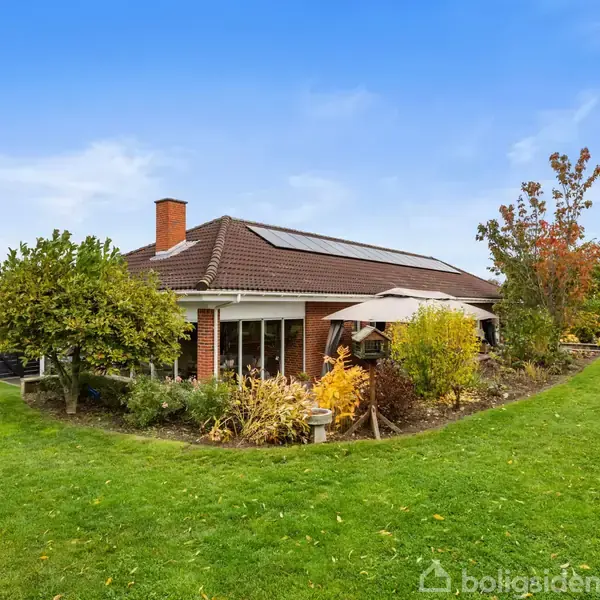 En etplans murstensvilla med solpaneler på taget står i en velplejet have med grønne plæner og træer, omringet af buske og en blå himmel i baggrunden.