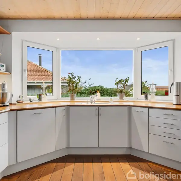 Et lyst køkken med hvidt interiør og trægulv, hvor planter pryder en stor vindueskarm. Udenfor ses et hus med et rødt tag.