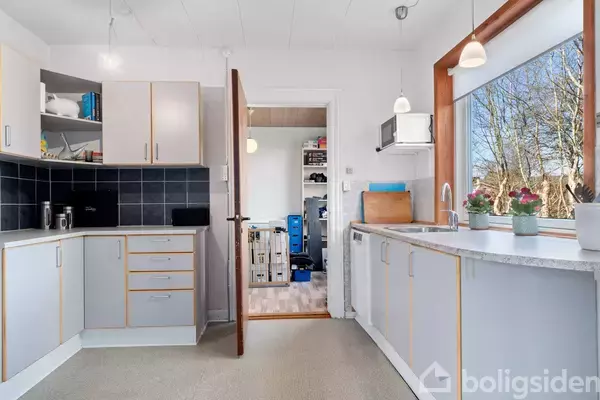 Et lyst køkken med moderne skabe og skuffer, en stor vask, og udsigt gennem et stort vindue. Køkkenet åbner ud til et rum med trægulv og opbevaringshylder.