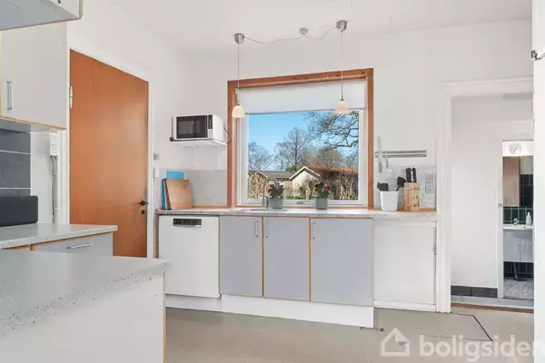 Køkken med lysegrå skabe, hvid bordplade og mikroovn på en hylde. Et stort vindue viser udsigt til have med træer. Rummet er lyst og skandinavisk i stilen.