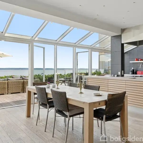 Moderne spisestue med lys trægulv og spisebord, omgivet af stole. Store vinduer åbner ud mod terrasse med udsigt over havet, hoftehøjt køkken i baggrunden. Ingen tekst til stede.