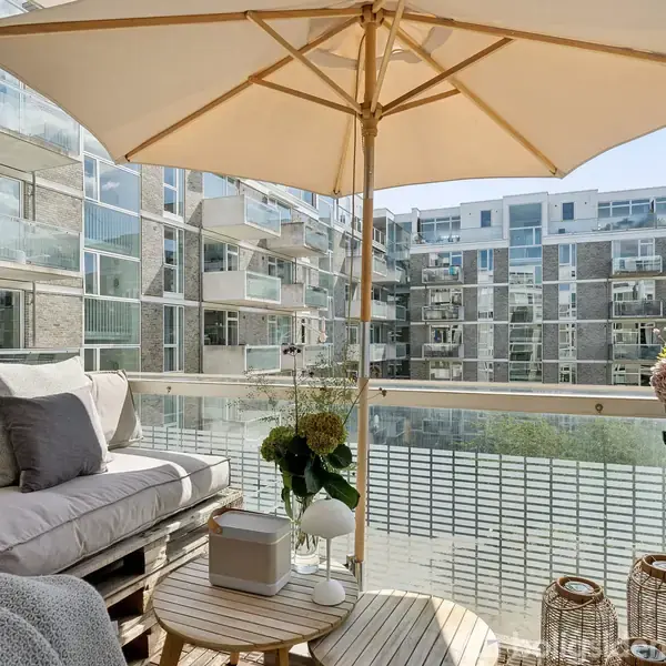 En altan med en stor parasol skygger over en grå sofa fyldt med puder. Små borde med planter og lanterner skaber en hyggelig stemning omgivet af moderne lejlighedsbygning.