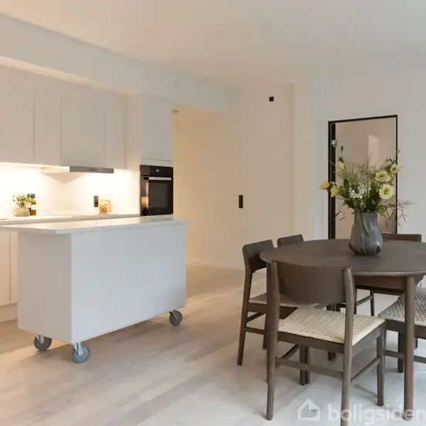 Køkkenområde med hvidt køkkenbord på hjul, omkranset af moderne, hvide skabe. Rundt træbord med fire stole, vase med blomster. Lys, minimalistisk indretning. Plant i hjørnet.