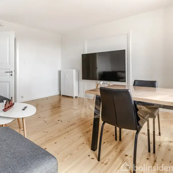 Stue med en sofa til venstre, et rundt sofabord med dekorationer, et spisebord til højre med tre stole, og et fladskærms-tv på væggen. Trægulv omgiver området.