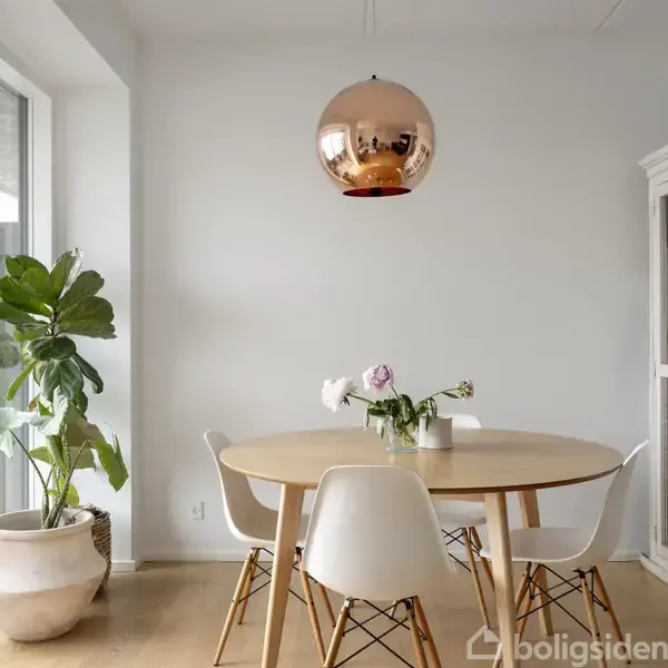 Rundt træbord omgivet af hvide stole står i lys spisestue. En kobberlampe hænger over bordet. Bagved ses en stor plante i hvid potte og et vitrineskab til højre.