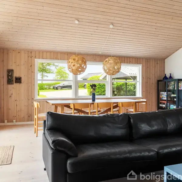 Sort lædersofa i en stue, står foran et stort spisebord med flerstole, placeret ved vinduer med udsigt til en græsplæne. To hængelamper hænger over bordet.