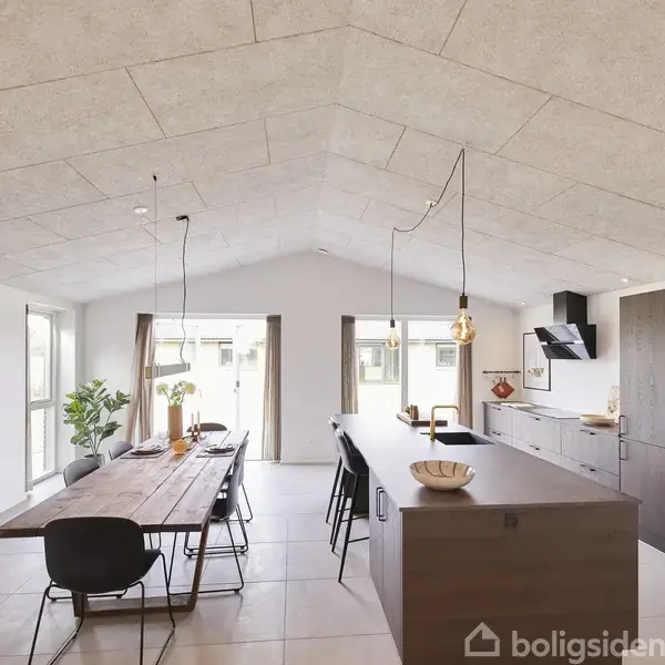 Et moderne køkken-alrum med et langt træbord omgivet af stole. Køkkenøen modsat har en indbygget vask. Store vinduer og minimalistisk indretning præger rummet.