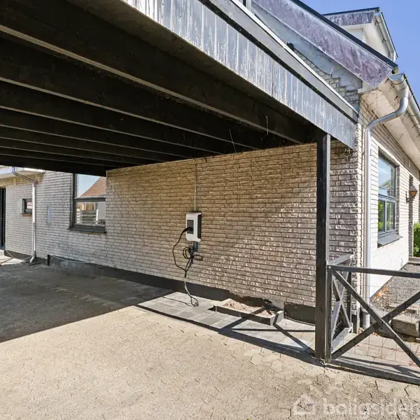 Carport med ladestik hænger fra hvid murstensvæg. Derudover ses en del af et ensidet hus og en stenterrasse. Omkringliggende haver med grøn beplantning under blå himmel.